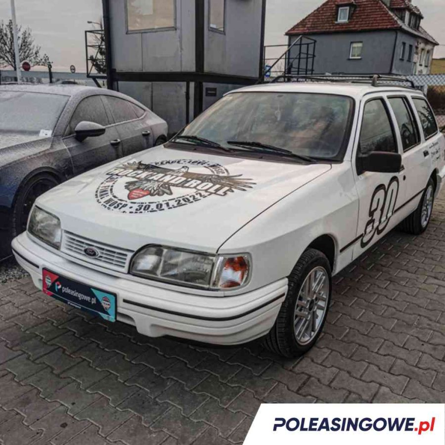 Biały samochód marki Ford Sierra z numerem startowym '30' i logotypem 'Rock and Roll' zaparkowany na placu przed budynkiem. Na pojeździe widoczne są naklejki sponsorów, w tym Poleasingowe.pl.