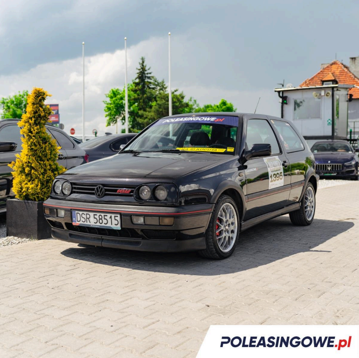 Czarny Volkswagen Golf III GTI z numerem rejestracyjnym DSR 58515, wystawiony na sprzedaż przez Poleasingowe.pl. Samochód stoi na parkingu, otoczony zielenią i budynkami, a na szybie widnieje naklejka Poleasingowe.pl. Na bocznej części samochodu znajdują się dodatkowe oznaczenia, w tym numer 1395, sugerujące udział pojazdu w wydarzeniach motoryzacyjnych lub aukcji. To klasyczne auto sportowe, utrzymane w dobrym stanie, przyciąga uwagę jako oferta dla fanów motoryzacji.